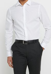 Camicia bianca con bottoni e maniche lunghe, abbinata a pantaloni grigio scuro e una cintura nera con fibbia argentata. Tessuto dalla texture liscia.
