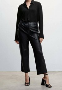 Femme portant une chemise noire boutonnée, un pantalon large en cuir noir court et des sandales noires à talons hauts avec des brides autour de la cheville.