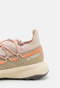 Beige och ljusrosa träningsskor med orange detaljer, texturerad ovandel, flexibla sula och elastiskt snörsystem. Varumärkeslogotyp synlig.