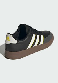 Crne tenisice s žutim i bijelim trakama, gumeni potplat i suede naglasci na prstima; imaju podstavljeni ovratnik i teksturiranu završnu obradu.