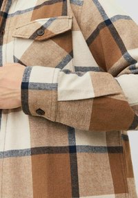 Bruine en crèmekleurige geruiten overhemd met een gestructureerde stof; heeft een knoopjes borstzak en opgerolde mouwen die contrasterende marineblauwe strepen tonen.