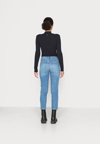 Persona con cabello rizado en un moño bajo, usando una camiseta negra de manga larga con canalé, jeans azul claro y botas negras hasta el tobillo, de pie frente a una pared blanca.