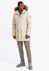 Beige péřová bunda s kožešinovou kapucí, dvěma předními kapsami a zipovým zapínáním. V kombinaci s svetle hnědými cargo kalhotami a hnědými šněrovacími botami.