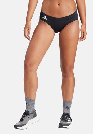 Mujer con bragas deportivas negras, calcetines grises Adidas y zapatillas de correr grises, de pie frente a un fondo blanco.