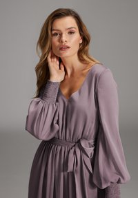 Leichter, fließender, lila Dress mit langen, gepufften Ärmeln und einem V-Ausschnitt. Hat eine geraffte Taille und eine gebundene Gürteldetails. Weicher Stoff.