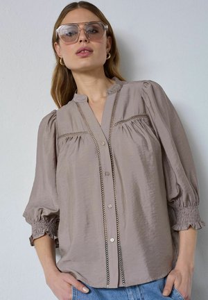 Frau mit übergroßen, durchsichtigen quadratischen Sonnenbrillen, goldenen Creolen, taupefarbener Bluse mit Spitzenbesatz und blauen Jeans, stehend vor einer weißen Wand.