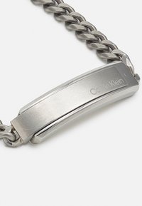 Sidabrinė metalinė grandinėlė su poliruotu stačiakampiu užsegimu. Užsegime įspaustas logotipas "Calvin Klein". Sklandi tekstūra, pramoninis dizainas.