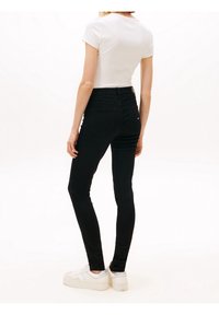 Woman standing wearing black skinny jeans, white short-sleeve top, and white sneakers against a plain white background.