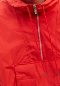 Röd vindjacka med hög krage, silverfärgad dragkedja och justerbara snoddar. Har en stor framficka och en mjuk nylontextur.