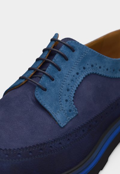 Chaussure habillée à bout golf en daim bleu avec perforations brogue, lacets bleu marine et panneau contrastant bleu plus clair près des œillets.