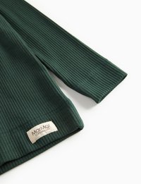 Tummanvihreä ribattuja pitkähihainen vaate, jonka helmassa on ommeltu valkoinen kangaslabel, jossa lukee "MarMar Copenhagen", valkoista taustaa vasten.