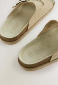 Bež nubuk slides sandale s zakrivljenim dnom, s teksturiranom unutrašnjosti i svijetlosmešnim gumastim đonom. Minimalistički dizajn, otvoreni prst.