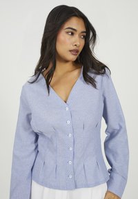 Donna con lunghi capelli scuri che indossa una camicetta a righe sottili blu chiaro, con bottoni e maniche lunghe, che guarda a destra contro uno sfondo neutro.