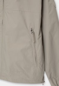 Veste gris clair en matériau lisse et léger, avec une poche zippée sur le côté gauche et une coupe décontractée. Coutures visibles.