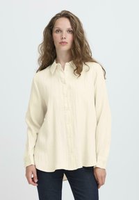 Woman with long wavy hair wears a loose, cream-colored button-up shirt and dark jeans, standing against a plain light background.