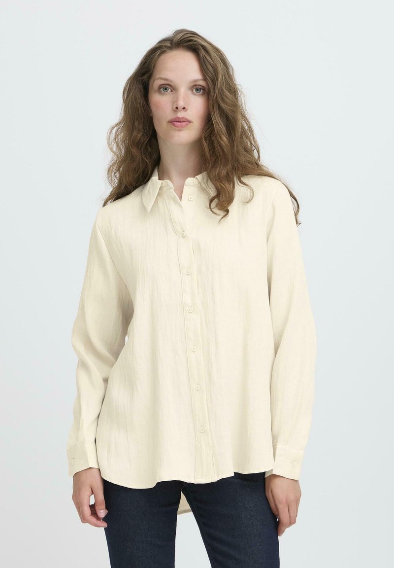 Woman with long wavy hair wears a loose, cream-colored button-up shirt and dark jeans, standing against a plain light background.
