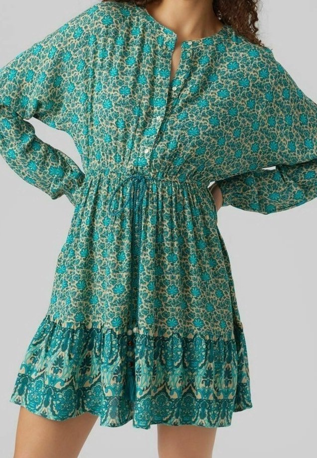 Femme portant une robe à manches longues à motif floral avec taille froncée et ourlet volanté dans des tons de bleu sarcelle et crème.