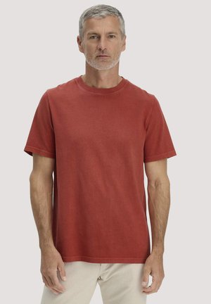 Mittelalterlicher Mann mit grauem Haar und Bart, der ein einfarbiges rotes T-Shirt mit kurzen Ärmeln und helle Hosen trägt, vor einem neutralen Hintergrund steht.