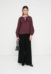 Bordeaux-top met gerimpelde accenten en lange mouwen, gecombineerd met brede zwarte broek. Model houdt een kleine zwarte handtas vast en staat tegen een effen achtergrond.