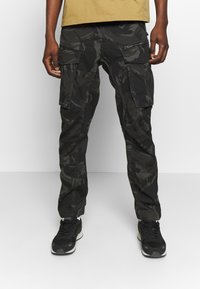 Man draagt zwarte en donkergrijze camouflage cargobroek met meerdere zakken en zwarte sneakers, op een effen achtergrond.