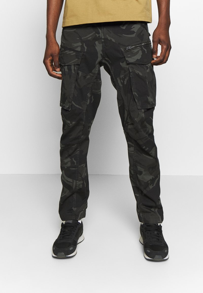 Man draagt zwarte en donkergrijze camouflage cargobroek met meerdere zakken en zwarte sneakers, op een effen achtergrond.