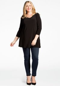 Schwarze, knielange Tunika mit drei Viertelärmeln, kombiniert mit dunklen Skinny Jeans und schwarzen High Heels. Angenehme Textur, lässiges Design.