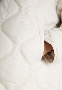 Veste matelassée beige clair avec une surface texturée, présentant une main visible dans la poche latérale et des motifs de couture courbes.