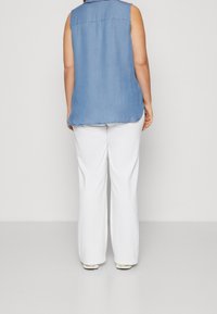 Slibetop i denim uden ærmer i lyseblå, med rygstykke og buet kant, parret med hvide brede bukser. Glat tekstur uden mønstre.