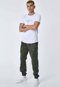 Giovane uomo con capelli biondi corti, in piedi con le braccia incrociate, indossa una maglietta bianca a maniche corte, pantaloni cargo oliva e sneakers bianche su sfondo bianco.