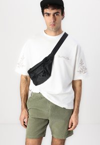 Schwarze Nylon-Crossbody-Tasche mit einem Reißverschlusstasche, getragen über einem weißen T-Shirt mit leicht floralem Stickdesign, kombiniert mit grünen Cordshorts.