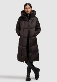 Schwarzer Steppmantel mit hohem Kragen, übergroßer Kapuze, gestepptem Design und Frontreißverschluss. Kombiniert mit schwarzen kniehohen Stiefeln.