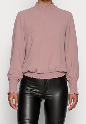 Długa bluzka w kolorze wrzosowym z falbanami na wysokim kołnierzu i mankietach, zestawiona z czarnymi dopasowanymi spodniami ze skóry.