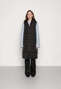 Melna polsterēta veste bez piedurknēm ar priekšpuses rāvējslēdzēju un horizontālām šuvēm. Valde virs gaiši zilas garumu piedurkņu blūzes.