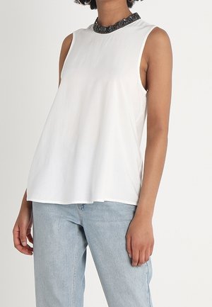 Kvinde iført hvid ærmeløs top med sort blondekrave og lyseblå jeans, stående med den ene arm afslappet ned langs siden.