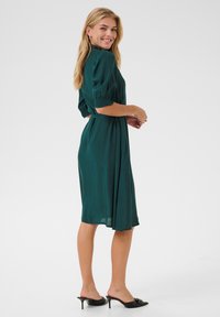 Abito turchese in tessuto leggero, con maniche a sbuffo corte, vita cintrata, gonna di lunghezza media e silhouette fluida. Mule nere con tacco.
