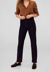 Jean en denim foncé taille haute avec surpiqûres contrastées, associé à une chemise marron boutonnée et des mocassins noirs. Fond uni.