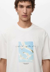 Hvid bomulds t-shirt med et grafisk print af hvide måger mod en lyseblå baggrund, med tekst under billedet i sort.