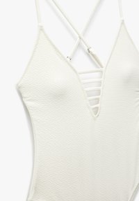 Maillot de bain blanc en tissu texturé à motif en diamant. Doté d'un décolleté en V profond et de bretelles croisées dans le dos.