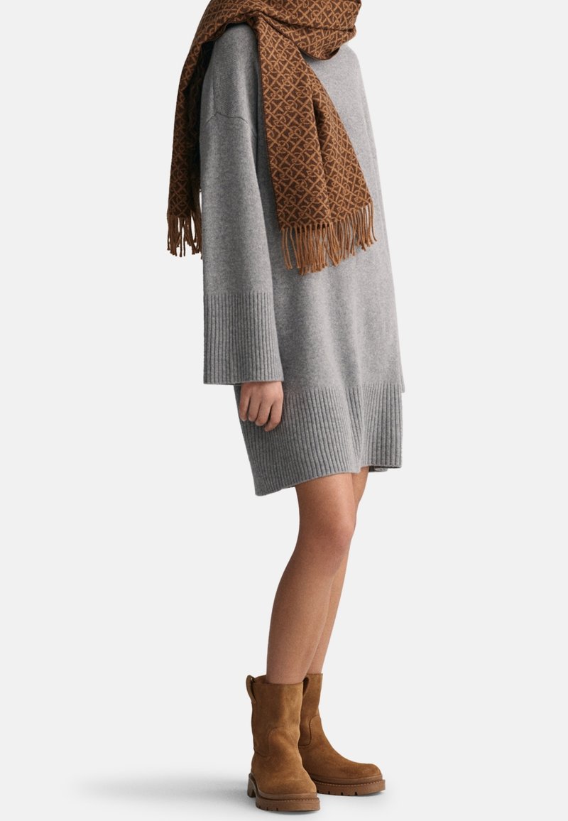 Personne portant une robe pull longue grise, une écharpe marron à motifs et des bottines en suède marron sur fond blanc uni.