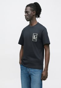 Νέος άντρας με σκουρόχρωμη επιδερμίδα και dreadlocks, που φοράει μαύρο μπλουζάκι με λευκό ορθογώνιο λογότυπο στο στήθος και μπλε τζιν, στέκεται πλάγια.