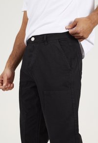 Pantalones cargo negros fabricados con tejido duradero, que cuentan con bolsillos laterales y cierre de botón. El diseño incluye una pierna recta y costuras mínimas.