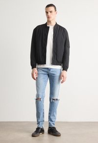 Svart bomberjakke med ribbestrikkede mansjetter, paret med lyseblå ripped jeans og svarte joggesko. Modellen står mot en nøytral bakgrunn.