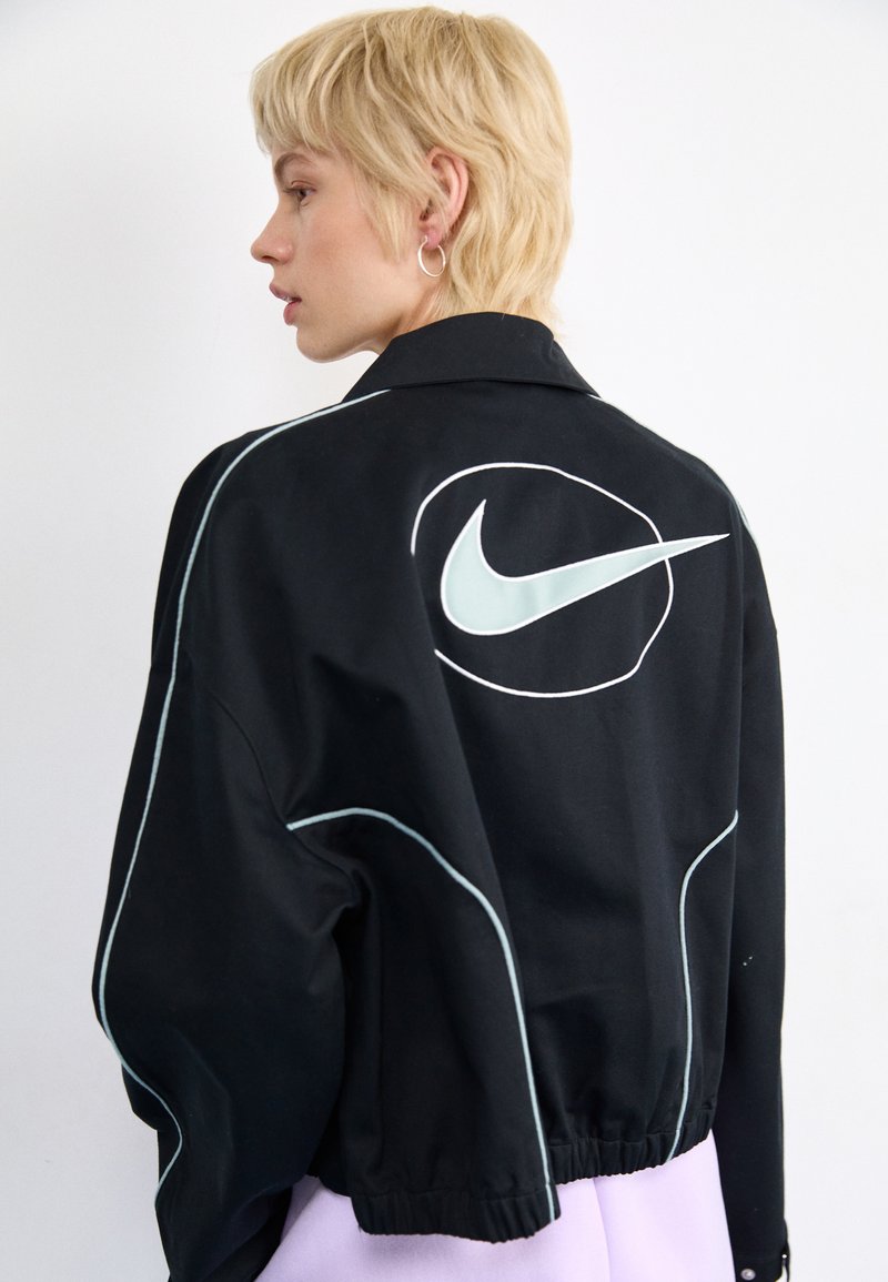 Chaqueta negra de Nike con costuras de acento blancas y un gran logo en la parte posterior. Cuenta con un cuello y un dobladillo elástico.