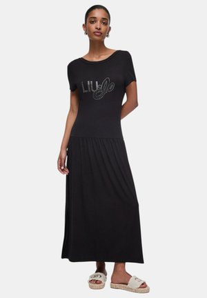 Donna che indossa un abito lungo nero a maniche corte con la scritta "LIU·JO" e sandali bianchi con suola intrecciata, posando davanti a uno sfondo bianco.