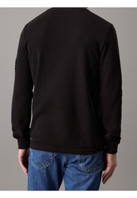 Black long-sleeve shirt with a textured, ribbed design. Features a rounded neckline and fitted cuffs, worn with blue denim jeans.