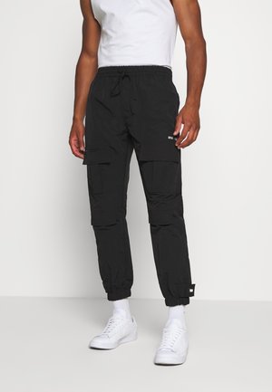 Homme portant un pantalon cargo noir avec plusieurs poches, des poignets élastiques, des chaussettes blanches, des baskets blanches et un débardeur blanc, debout sur un sol gris.