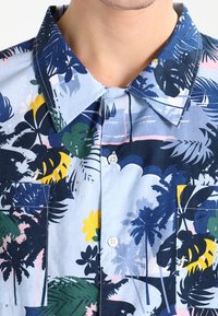 Kurzes, ärmelloses Hemd mit einem hellblauen Hintergrund, das ein tropisches Design mit grünen und gelben Blättern, navyblauen Akzenten und zwei Brusttaschen aufweist.