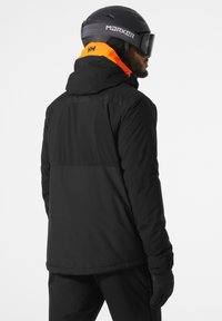Veste de ski noire avec des accents orange, dotée d'une capuche et d'un design épuré. Les gants sont sombres, complémentant l'ensemble de la tenue.