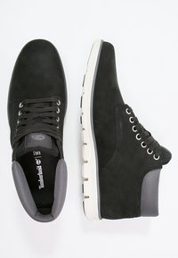 Svarta mockaskor med vita gummisålar, med runda snörningar, en vadderad krage och en strukturerad tygdetalj i grått.