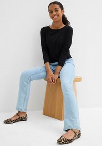 Top nero a maniche lunghe, jeans dritti blu chiaro e ballerine con stampa leopardata. Seduta su uno sgabello di legno contro uno sfondo semplice.